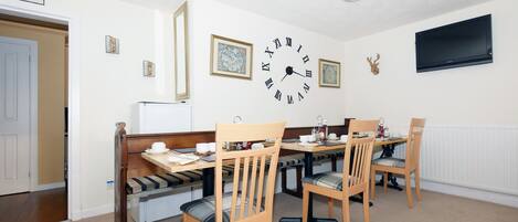 Uma área de refeições com uma mesa e cadeiras de madeira, um relógio de parede e uma televisão fixada na parede.