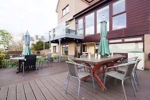 Una terraza de madera con mesa y sillas, barandilla de cristal y una casa de fachada marrón con ventanas amplias.