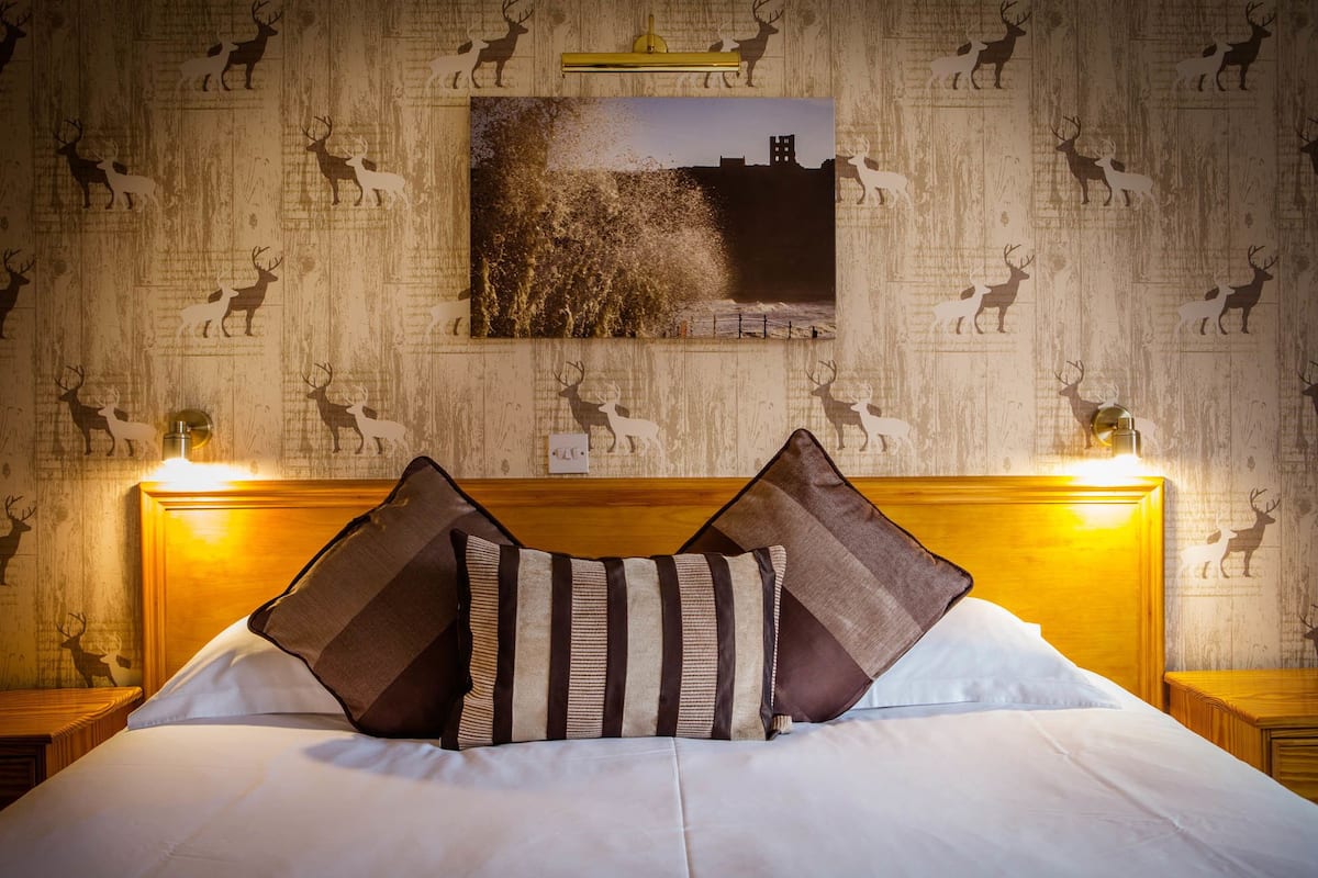 Quarto de hotel com cama, duas mesas de cabeceira, parede com estampas de veados e um quadro emoldurado sobre a cama.