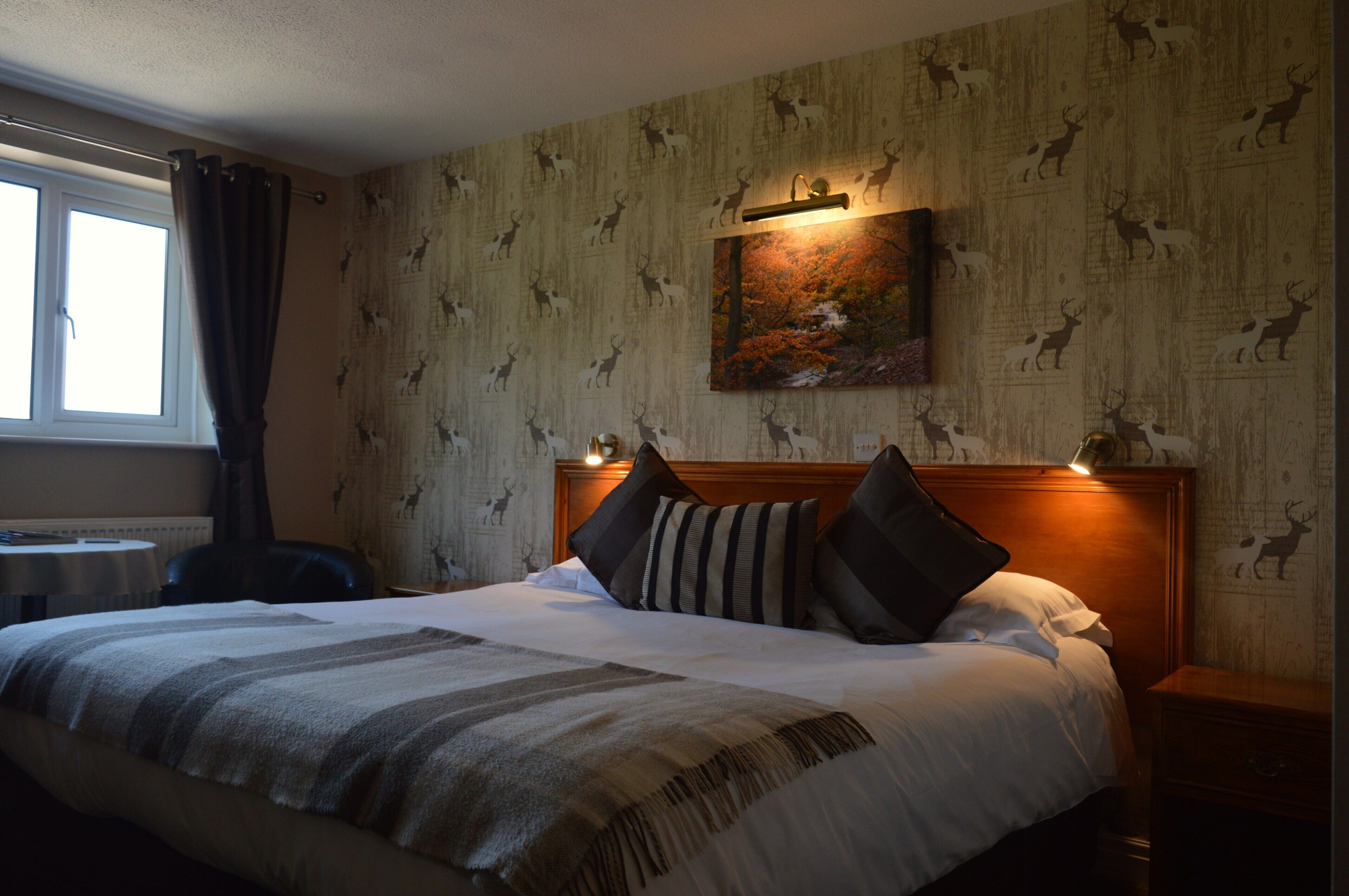 A hotel room with a large bed, a headboard with lights, a painting of deer on the wall, and a window with curtains.
