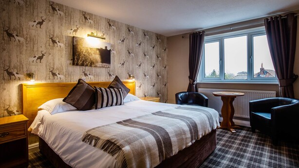 A hotel room with a large bed, a wooden headboard, a nightstand, a small table, a chair, and a window with curtains. - Orchard Lodge & Wolds Restaurant (Scarborough)