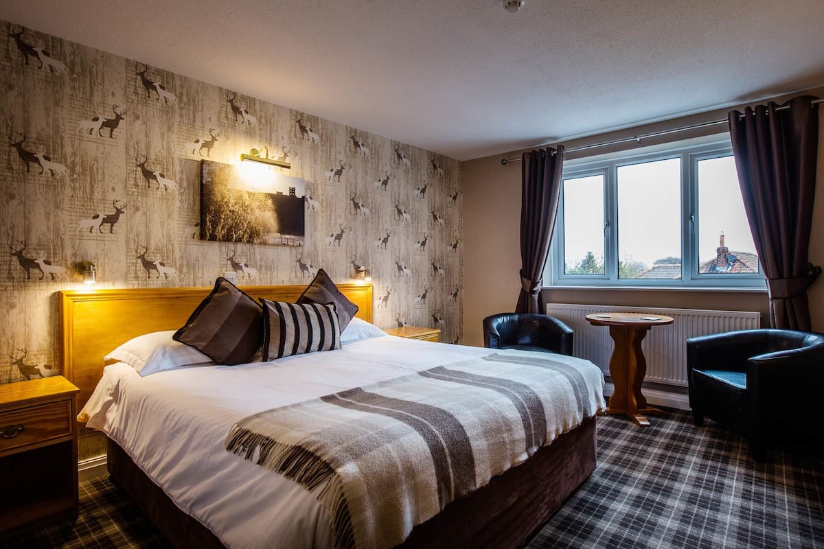 Quarto de hotel com uma cama grande, cabeceira de madeira, mesa de cabeceira, uma pequena mesa, uma cadeira e uma janela com cortinas.