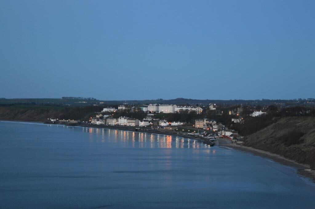 A coastal town with buildings along the waterfront, a clear sky, and calm water reflecting the buildings.