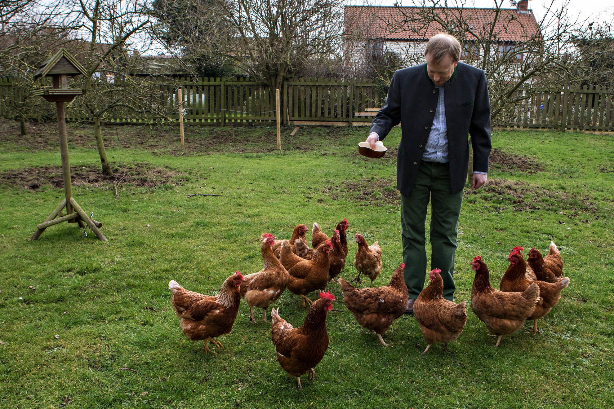 A man feeding chickens in a grassy area with a wooden fence and a birdhouse in the background.