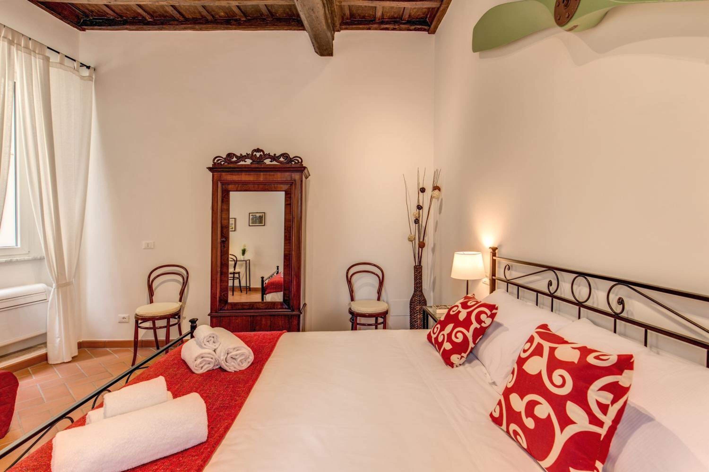 Quarto de hotel com cama, duas cadeiras, espelho, um vaso com plantas secas e janela com cortinas.
