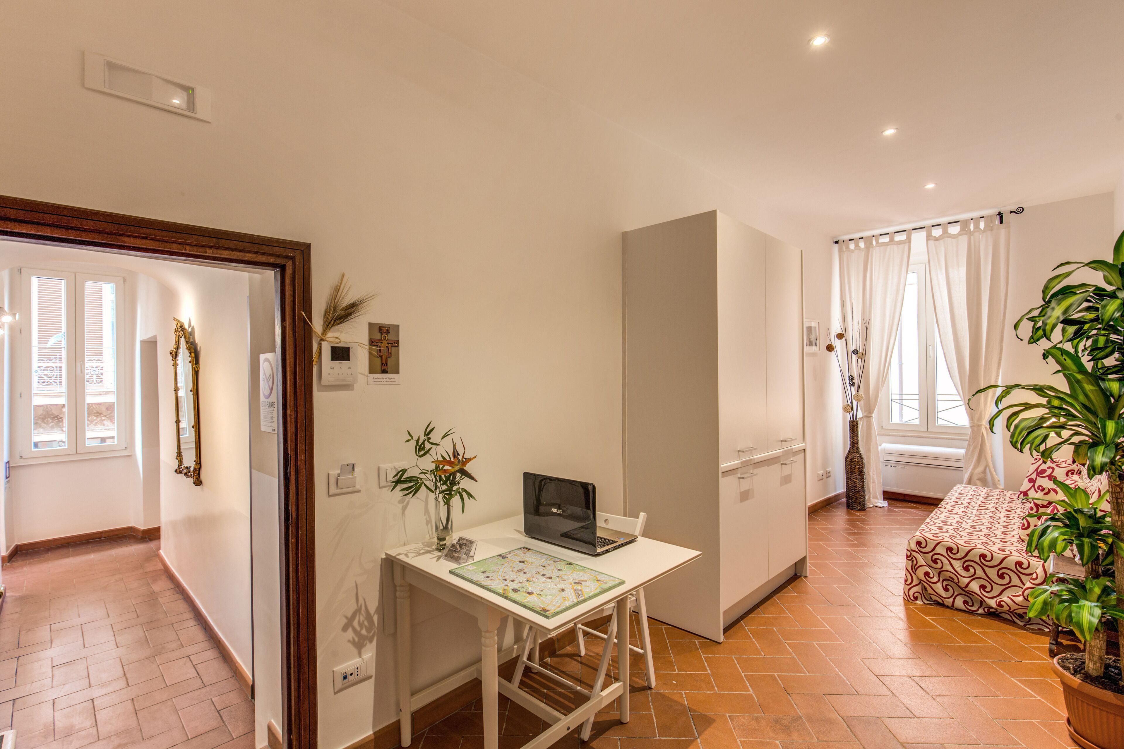 A room with a white desk, a laptop, a vase with plants, a bed with patterned bedding, and a window with curtains.