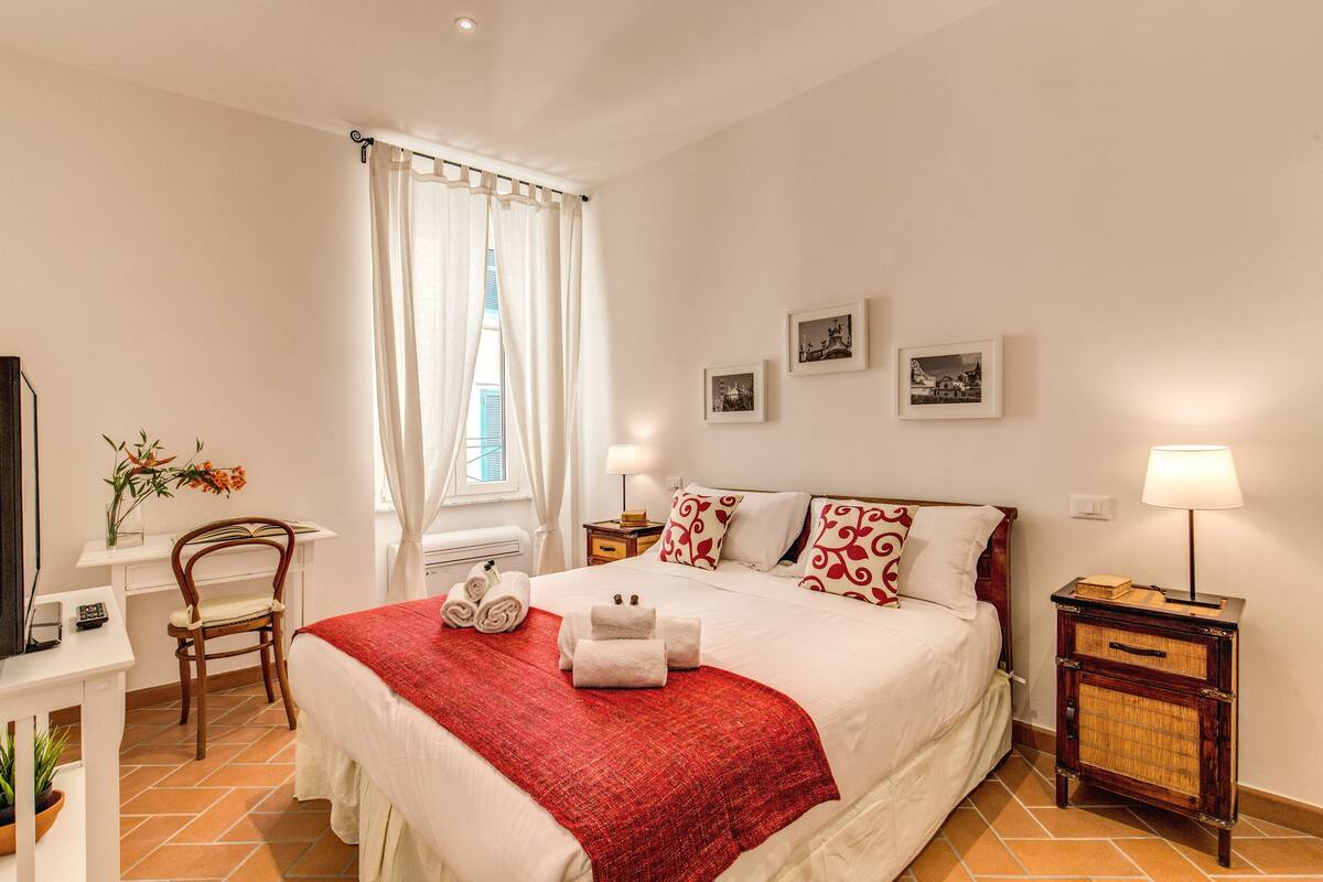 Habitación de hotel ordenada con cama, escritorio, silla, mesita de noche con lámpara y ventana con cortinas.