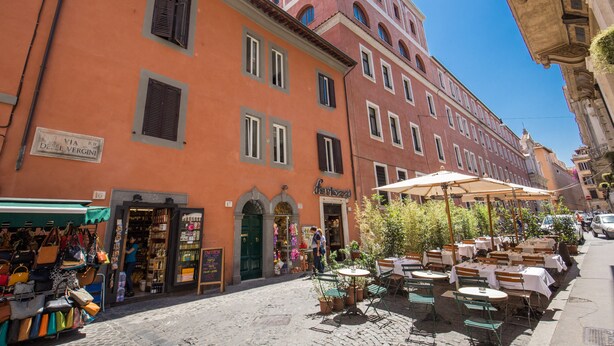 A cobblestone street with outdoor seating, a shop selling bags, and a building with a sign reading "ferragosto". - Piccolo Trevi Suites (Rome)