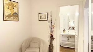 A room with a beige armchair, a decorative vase with branches, a framed picture, and a tiled floor. - Piccolo Trevi Suites (Rome)