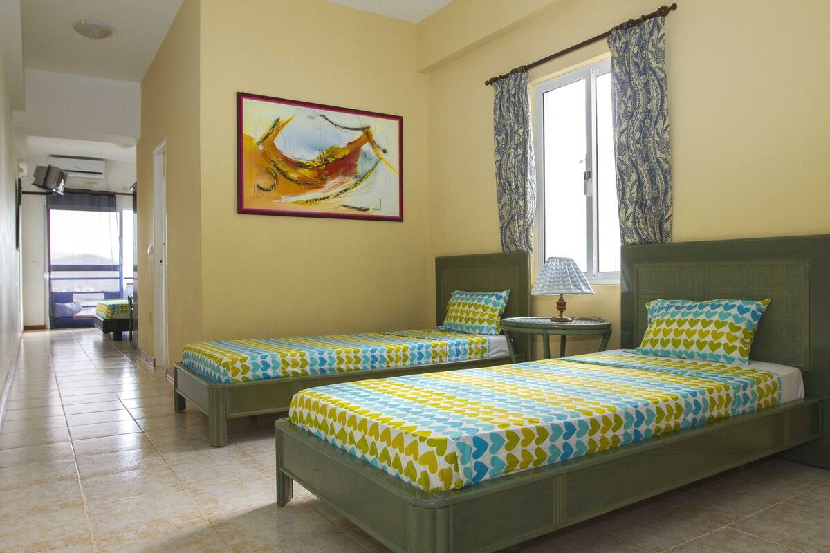 A hotel room with two single beds, a painting on the wall, a window with patterned curtains, and a view of the sea through the door.