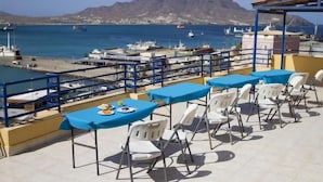 Um terraço com mesas e cadeiras com vista para uma marina, com barcos e uma montanha ao fundo.