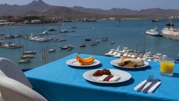 Mesa com itens de café da manhã e vista para uma marina com barcos e montanhas ao fundo.