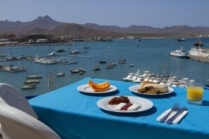 Ein Tisch mit Frühstücksangeboten und Blick auf einen Yachthafen mit Booten und Bergen im Hintergrund.