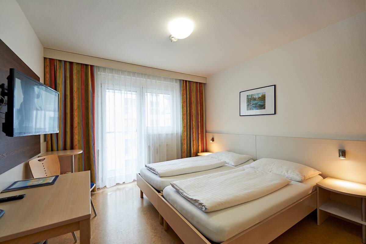 Ein Hotelzimmer mit zwei Betten, einem Schreibtisch, einem Fernseher und einem Vorhangfenster.