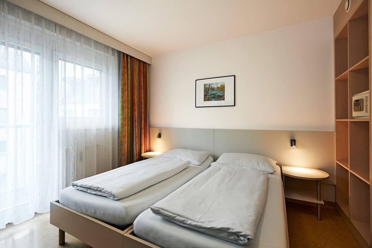 Quarto de hotel com duas camas, uma janela grande com cortinas translúcidas, um quadro na parede e uma mesa de cabeceira com abajur.