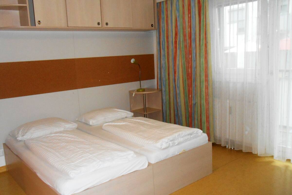 Ein Doppelbett mit weißer Bettwäsche in einem Zimmer mit Holzboden und einem gestreiften Vorhang.