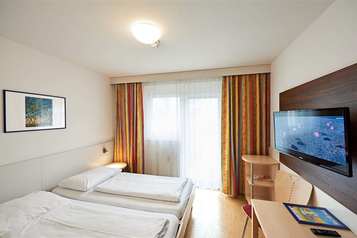 Une chambre d’hôtel avec deux lits, une télévision, un tableau et une fenêtre avec des rideaux.