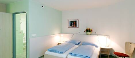 Quarto de hotel com uma cama, mesas de cabeceira, uma cadeira, uma prateleira na parede com flores e uma porta para o banheiro.
