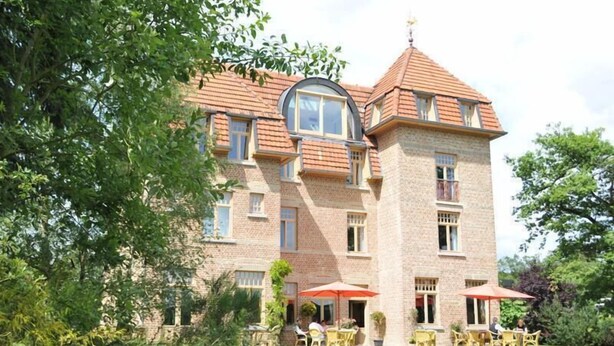 Un edificio grande de ladrillo con una torre destacada, múltiples ventanas y mesas exteriores con sombrillas.