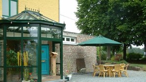 Un invernadero con puerta verde, pared de ladrillo y un patio con muebles de mimbre y un paraguas verde.