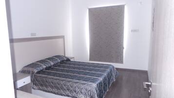 Een kleine, verzorgde slaapkamer met een bed, nachtkastje en raam met een patroonblik.