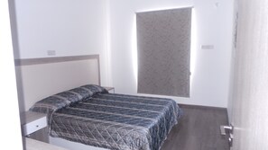 A small, neatly arranged bedroom with a bed, a bedside table, and a window with a patterned blind. - Neptune Hotel Apartments (Pegeia)