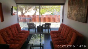 A room with two red sofas, a glass-top table, and a view of a red fence and trees through a sliding glass door. - Neptune Hotel Apartments (Pegeia)