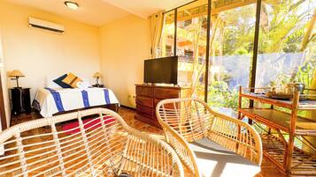 Una habitación con una cama, un televisor sobre un mueble de madera, sillas de mimbre y vistas a un jardín exuberante.