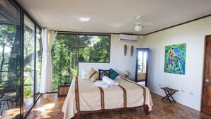 Un dormitorio con una cama grande, un ventilador de techo, una ventana con cortinas y vistas a árboles.