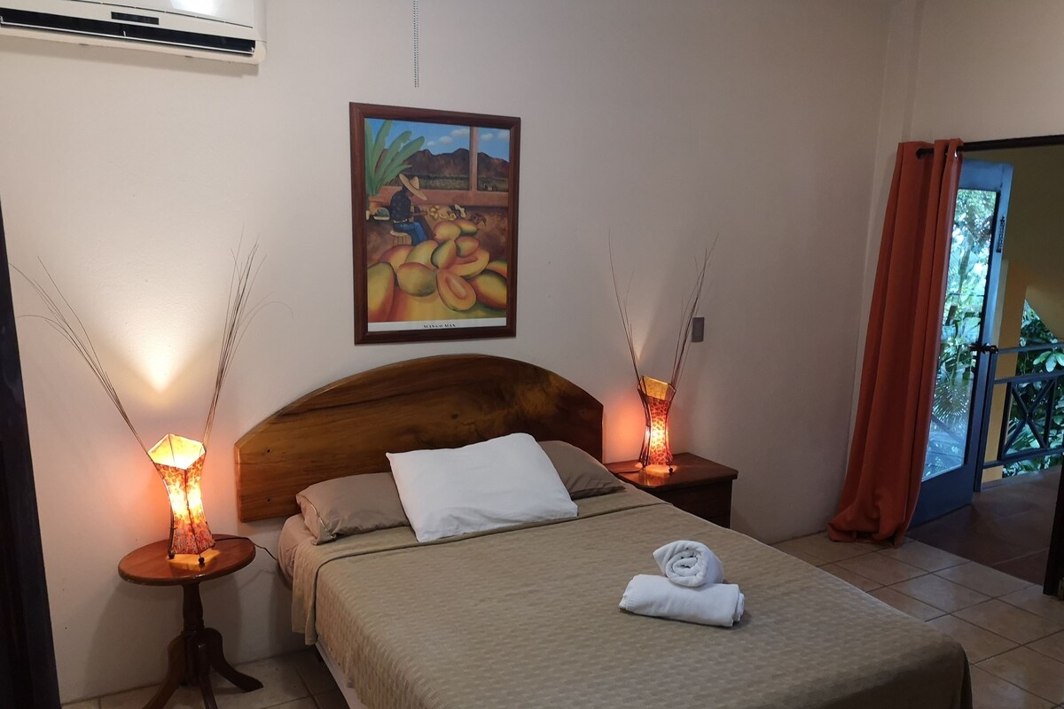 A bedroom with a wooden headboard, a bed with a beige bedspread, two bedside tables with lamps, a framed picture on the wall, and a view of greenery through a door.
