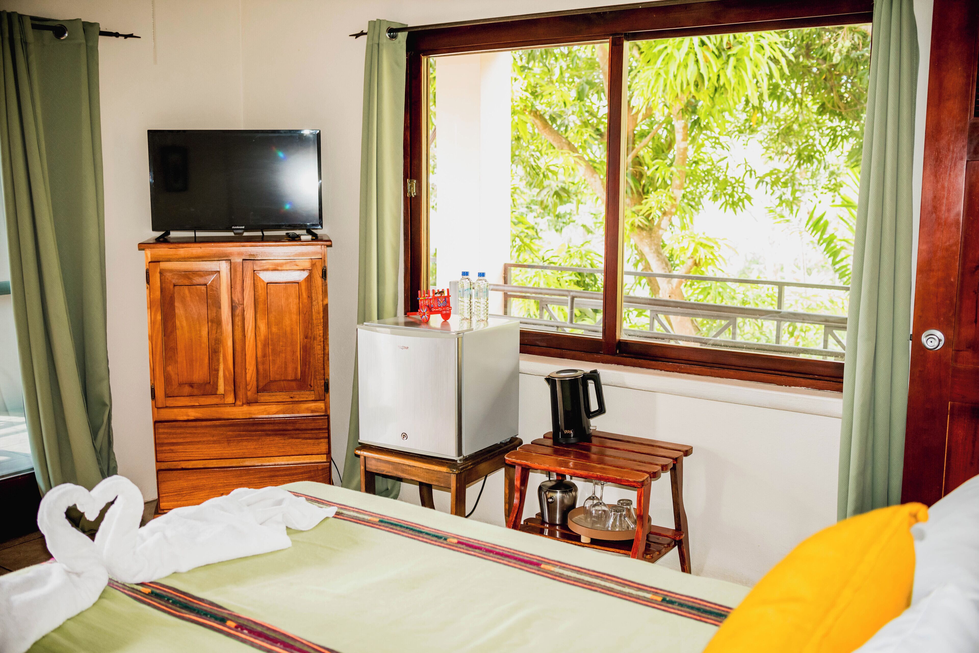 Une chambre d’hôtel avec un lit, un téléviseur posé sur une commode en bois, un petit réfrigérateur, une machine à café et une fenêtre donnant sur des arbres.