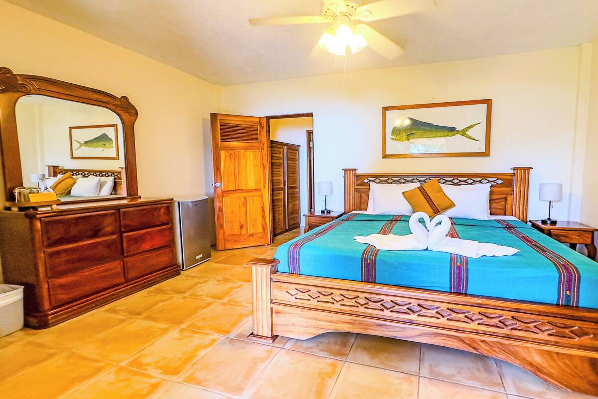Une chambre d’hôtel avec un lit, des meubles en bois, un ventilateur de plafond, un miroir et un tableau représentant des poissons accroché au mur.