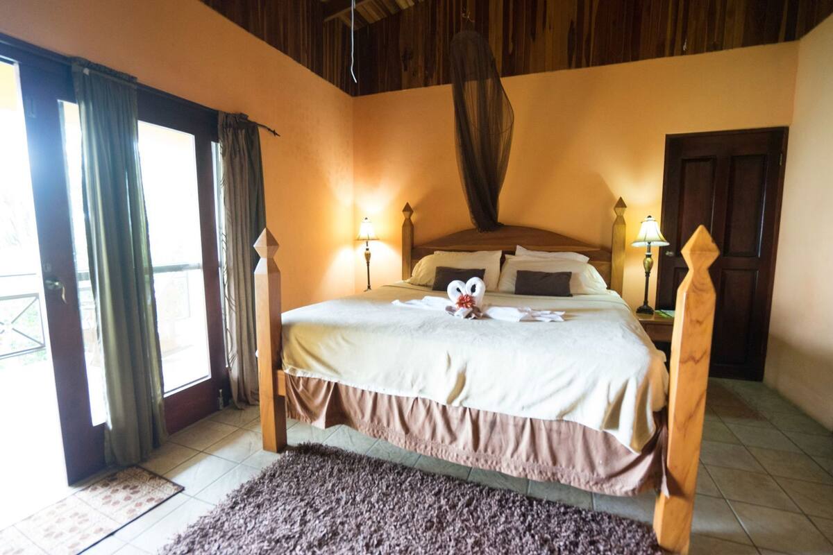 Une chambre à coucher avec un lit, des rideaux, une tête de lit en bois, une table de chevet et une lampe.