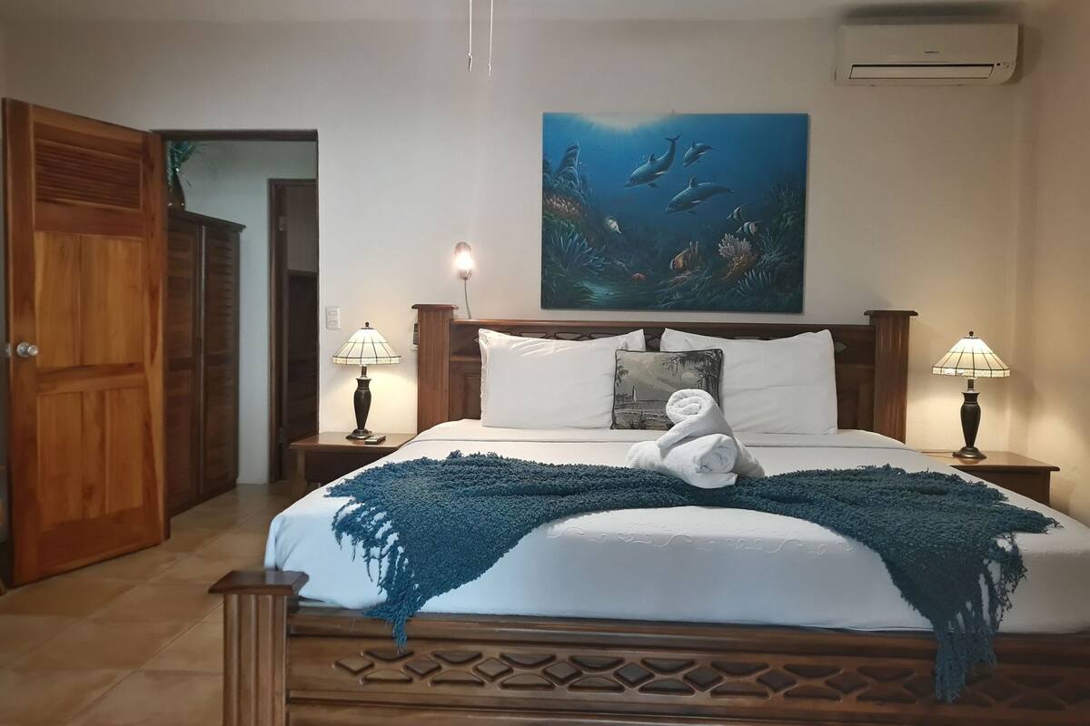 Une chambre d’hôtel avec un lit, deux lampes de chevet, un tableau représentant des dauphins et un appareil de climatisation.