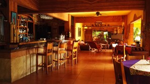 Zona de bar con mobiliario de madera, una barra con botellas y una zona de comedor con sillas y una mesa.