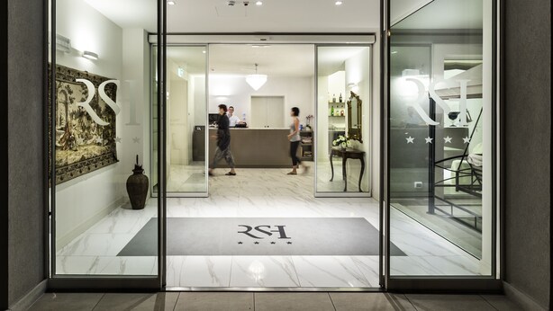 Entrada de hotel con mostrador de recepción, una recepcionista y una zona de espera con un espejo grande y elementos decorativos.