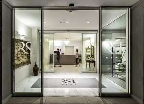 Entrada de hotel con mostrador de recepción, una recepcionista y una zona de espera con un espejo grande y elementos decorativos.