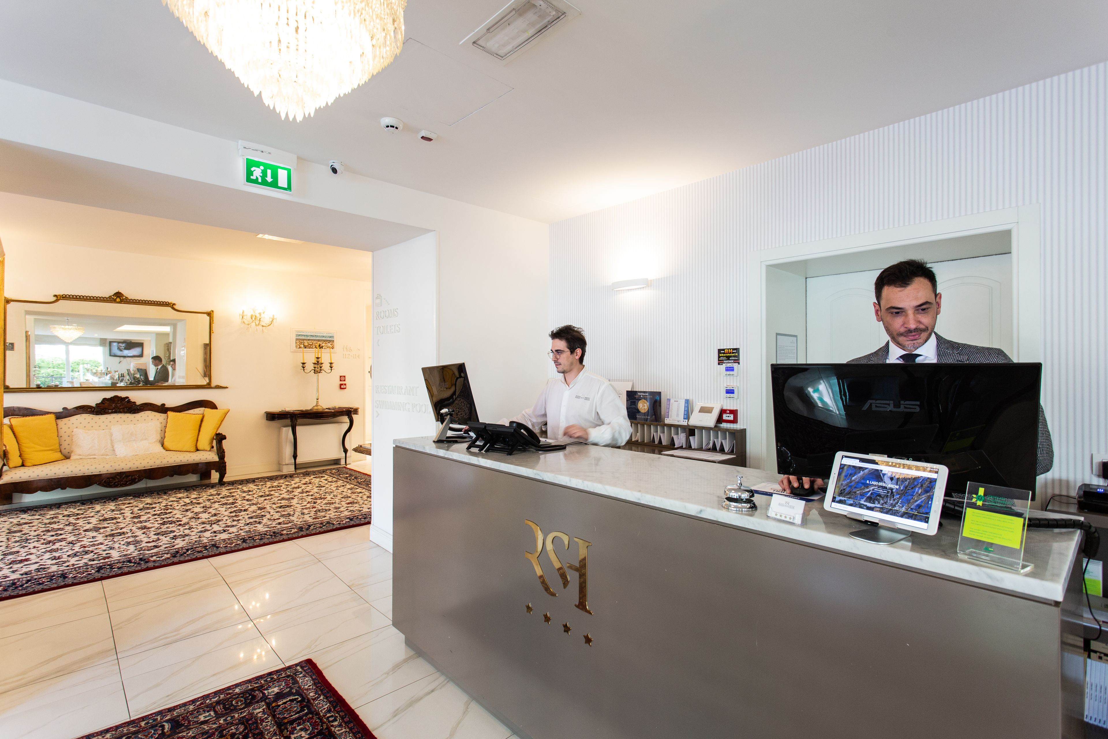 A hotel reception area with a reception desk, a sofa with yellow cushions, a chandelier, and a large mirror.