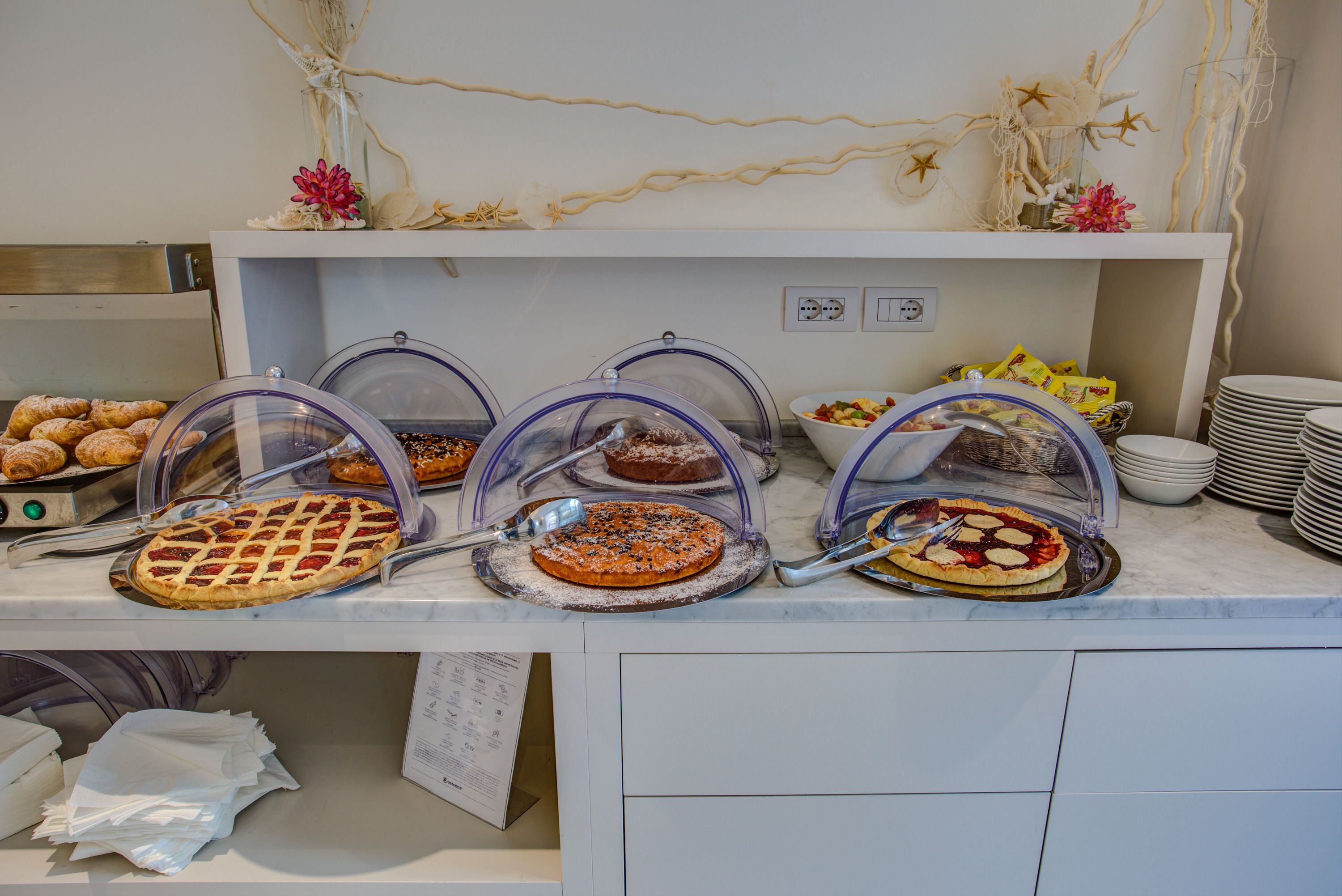 A buffet with various pies and pastries under protective covers, a bowl of fruit, and stacked plates.