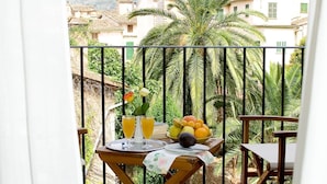 Un balcón con mesa lista para el desayuno, con frutas y bebidas, con vistas a una zona residencial con palmeras y edificios.