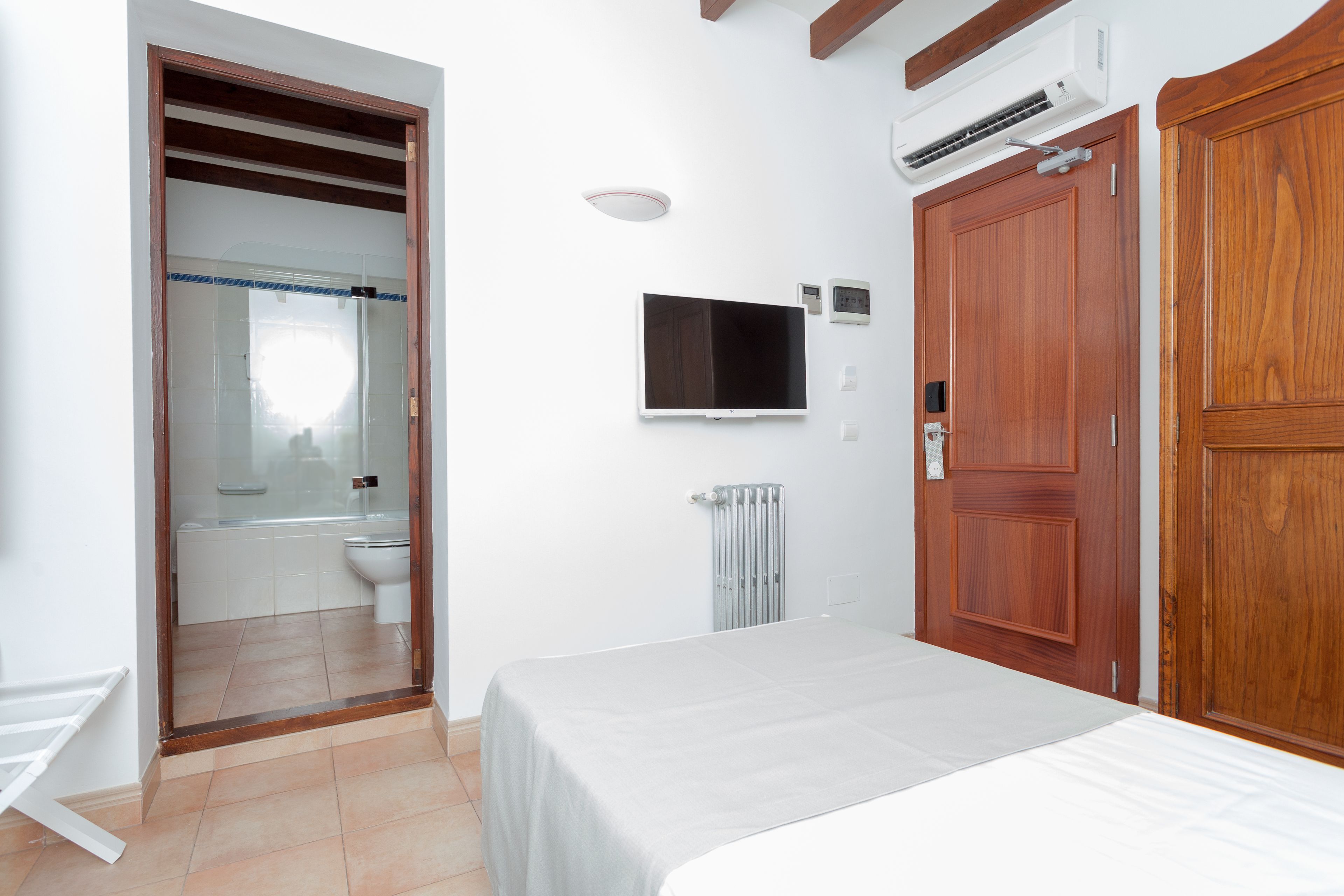 A bedroom with a bed, a wooden wardrobe, a TV mounted on the wall, and an air conditioning unit.