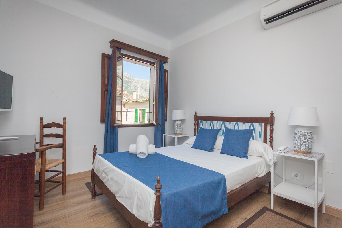 A bedroom with a wooden bed, a desk, a chair, a bedside table with a lamp, and a window with curtains.