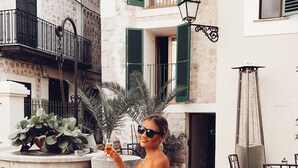 A woman seated at an outdoor table with a white tablecloth, surrounded by chairs and a potted plant. - Hotel El Guía (Sóller)