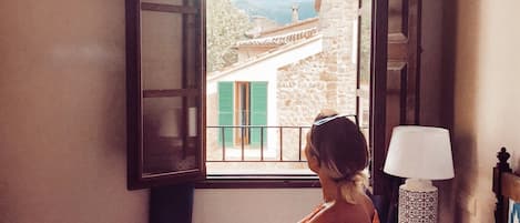 A person sitting on a bed in a room with a window, a bedside table with a lamp, and a view of a building outside.
