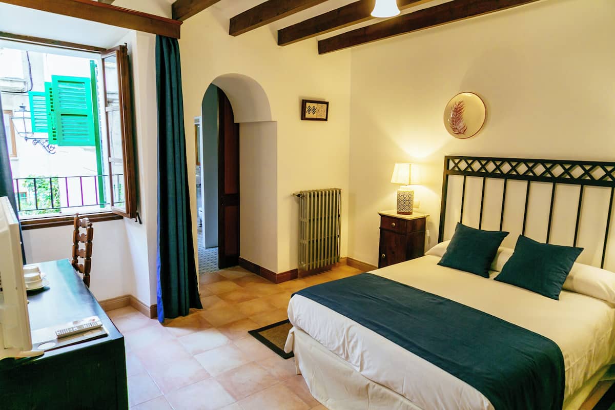 A bedroom with a bed, a desk, a window with curtains, a radiator, and a wall-mounted lamp.