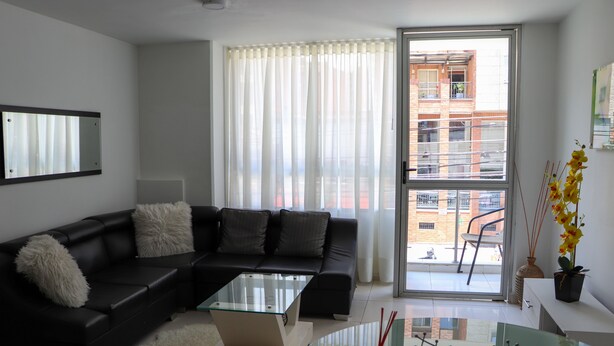 A modern living room with a black leather sofa, a glass coffee table, and a view of a cityscape through large windows. - Moblat Apartamentos (Medellín)