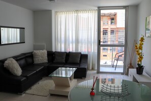 A modern living room with a black leather sofa, a glass coffee table, and a view of a cityscape through large windows. - Moblat Apartamentos (Medellín)