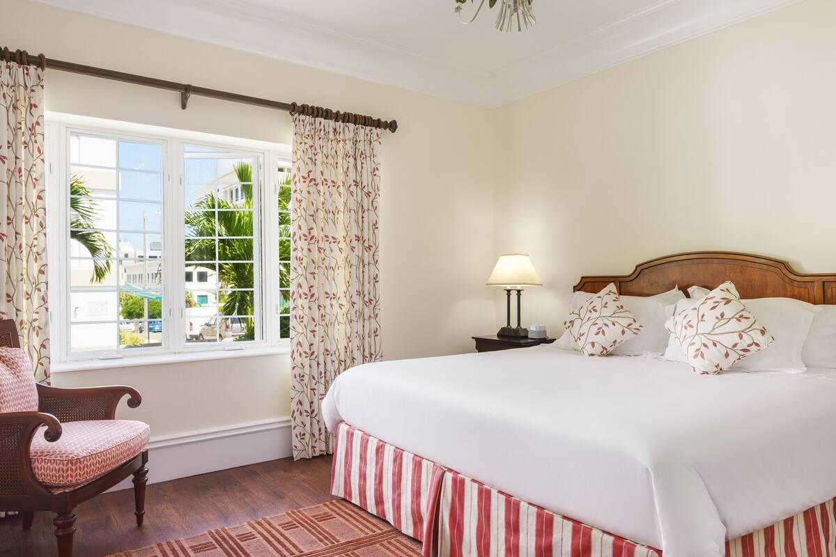 A bedroom with a large bed, a wooden headboard, a nightstand with a lamp, a chair, a window with curtains, and a chandelier.