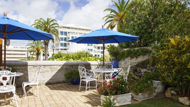 An outdoor patio with white metal chairs and tables, blue umbrellas, and potted plants. - The Oxford House (Pembroke Parish)