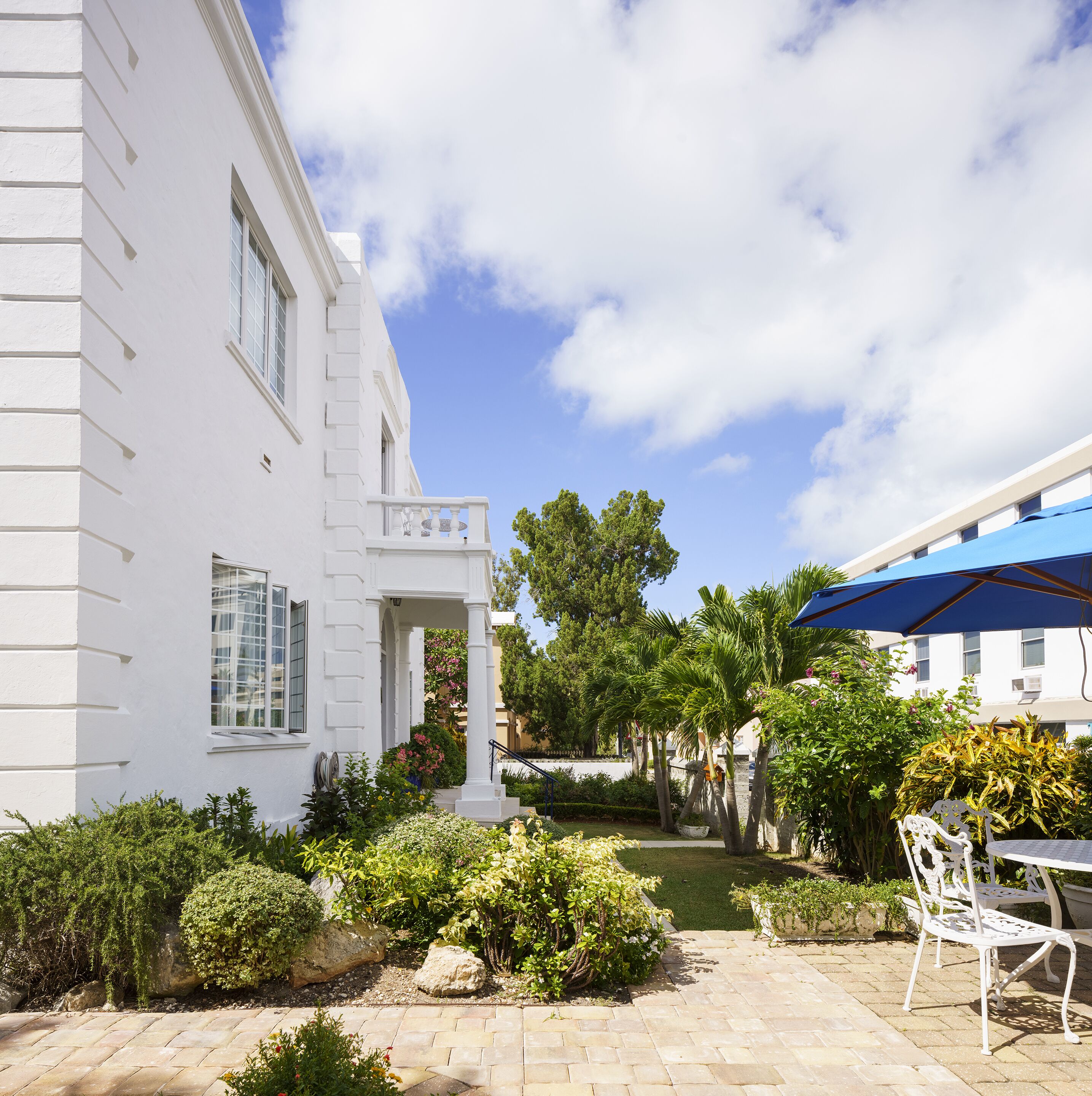 A white building with a balcony, surrounded by greenery and a paved patio area.